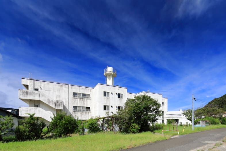 抜けるような青空を背景に建つ白いコンクリート造の建物。屋上に給水塔のような設備が見え、階段と緑に囲まれている